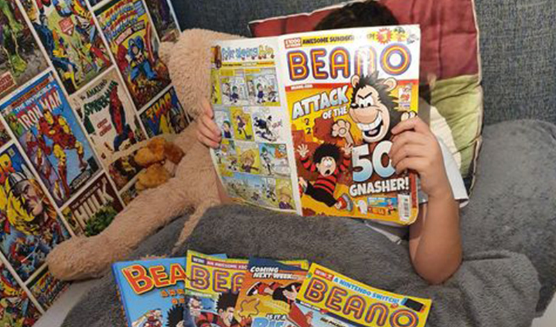 A child reading Beano magazine in bed with a selection of other comics on the covers.