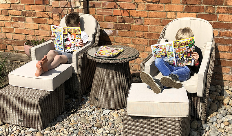 Two kids sat on garden furniture each reading a copy of the Beano comic; a pile of magazines is stacked between them